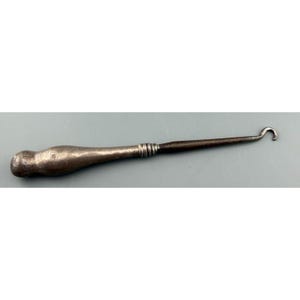 May include: An antique metal crochet hook with a silver-toned handle and a dark, slender hook. The handle has a bulbous end and a textured surface. The hook is a vintage tool.