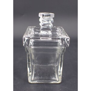 Puede incluir: Un tarro de vidrio transparente de forma rectangular con tapa a juego. La tapa tiene un asa cuadrada. El diseño es sencillo y elegante, con líneas limpias y una forma clásica. El vidrio es transparente, permitiendo ver el contenido.