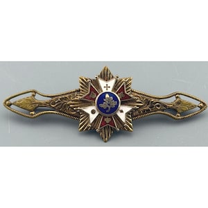 May include: An antique brooch with a starburst design. The brooch features a central blue circle with a leaf design, surrounded by red, white, and gold accents. The brooch has an ornate, elongated shape with decorative details.