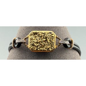 May include: A gold-toned bracelet with a rectangular, textured, gold-colored center stone. The bracelet features black leather straps and small, sparkling accents on the sides. The jewelry is displayed on a gray surface.