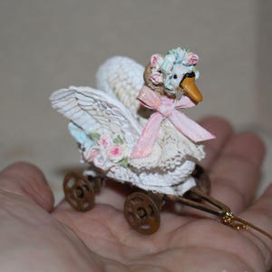 May include: A miniature swan figurine with a decorative carriage. The swan is white with detailed wings, a yellow beak, and a pink bow. It is adorned with floral accents and sits on a small, wheeled carriage.