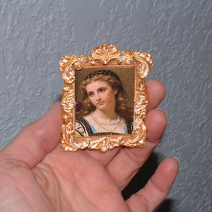 May include: A small, ornate gold-colored picture frame with a portrait of a woman. The frame has a detailed, scrollwork design. The portrait features a woman with long, wavy hair and a contemplative expression. The frame is approximately 2 inches by 2 inches.
