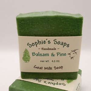 May include: Two bars of green goat milk soap stacked on top of each other. The top bar has a label that reads "Sophie's Soaps - Handmade - Balsam & Pine - Net WT. 4.5 OZ - Goat Milk Soap".