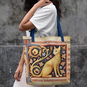 May include: Tote bag featuring a golden cat illustration in a medieval style. The bag has blue handles and a cream-colored background with ornate floral and star designs. The cat has blue eyes and is set within a red and blue border.