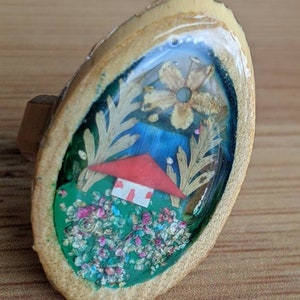 May include: A wooden ring with a clear resin dome containing a miniature house, flowers, and greenery. The house has a red roof and white walls. The flowers are pink, white, and yellow. The greenery is green and brown.