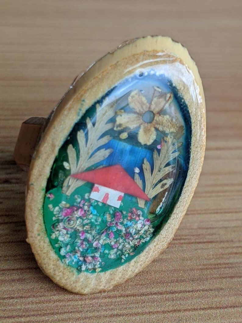 May include: A wooden ring with a clear resin dome containing a miniature house, flowers, and greenery. The house has a red roof and white walls. The flowers are pink, white, and yellow. The greenery is green and brown.