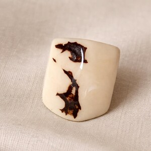 May include: A cream-colored, rectangular ring with dark brown, organic-shaped markings. The ring has a glossy finish and is set against a neutral fabric background. The design is abstract and naturalistic.