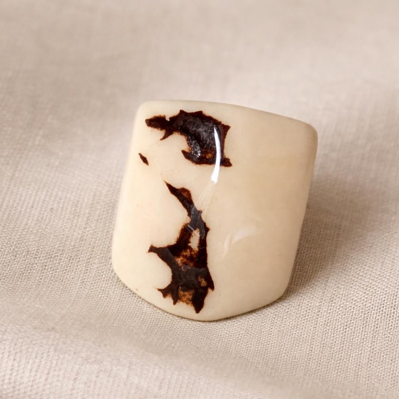 May include: A cream-colored, rectangular ring with dark brown, organic-shaped markings. The ring has a glossy finish and is set against a neutral fabric background. The design is abstract and naturalistic.