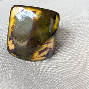 May include: A rectangular ring with a glossy finish. The ring features a blend of colors, including shades of green, yellow, and brown. The design has a marbled appearance, with the colors blending together. The ring is set against a neutral background.