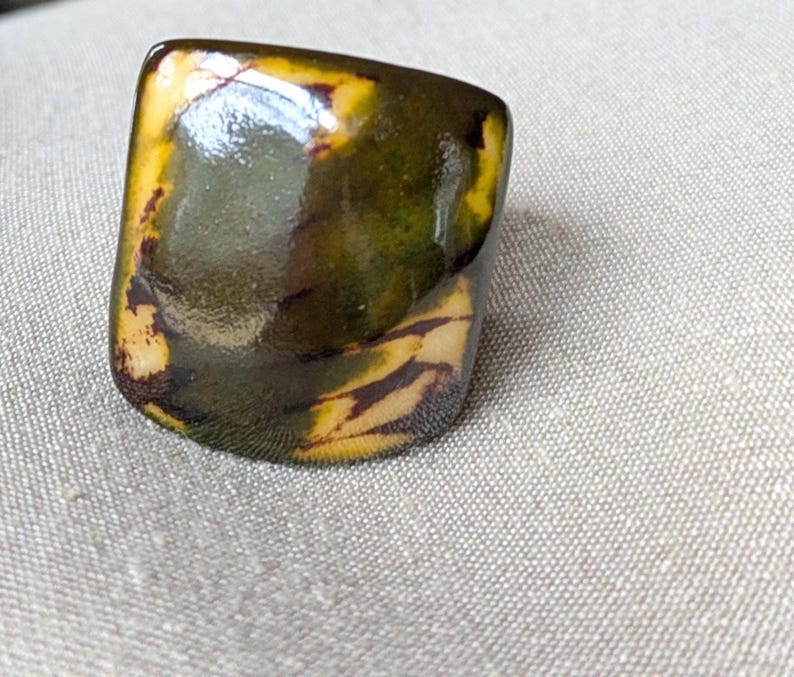 May include: A rectangular ring with a glossy finish. The ring features a blend of colors, including shades of green, yellow, and brown. The design has a marbled appearance, with the colors blending together. The ring is set against a neutral background.