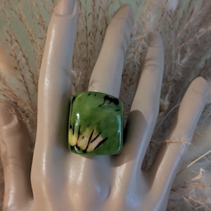 May include: A square-shaped ring with a green and yellow pattern. The ring is worn on a mannequin hand. The ring's design features a mix of green and yellow hues with black accents. The ring is a bold statement piece.