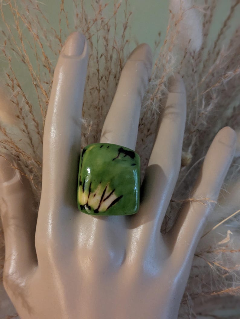 May include: A square-shaped ring with a green and yellow pattern. The ring is worn on a mannequin hand. The ring's design features a mix of green and yellow hues with black accents. The ring is a bold statement piece.