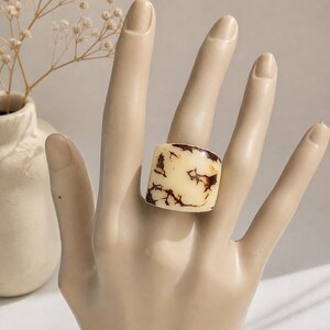 May include: A chunky, square-shaped ring with a cream-colored base and intricate brown veining. The ring is displayed on a mannequin hand, showcasing its bold design. The ring's unique pattern adds a touch of artistic flair to the jewelry.