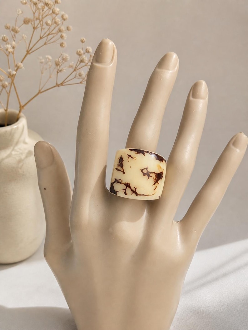 May include: A chunky, square-shaped ring with a cream-colored base and intricate brown veining. The ring is displayed on a mannequin hand, showcasing its bold design. The ring's unique pattern adds a touch of artistic flair to the jewelry.
