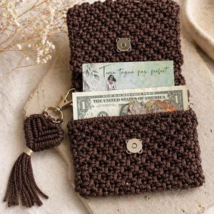 May include: A brown macrame wallet with a magnetic clasp, open to reveal a dollar bill and a card. The wallet has a matching tassel keychain. The card reads "Terra tagua paw perfect".