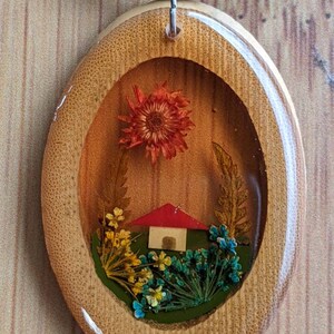 May include: A wooden oval pendant necklace with a clear resin insert featuring a miniature house and pressed flowers. The house has a red roof and white walls. The flowers are in shades of orange, yellow, blue, and green.