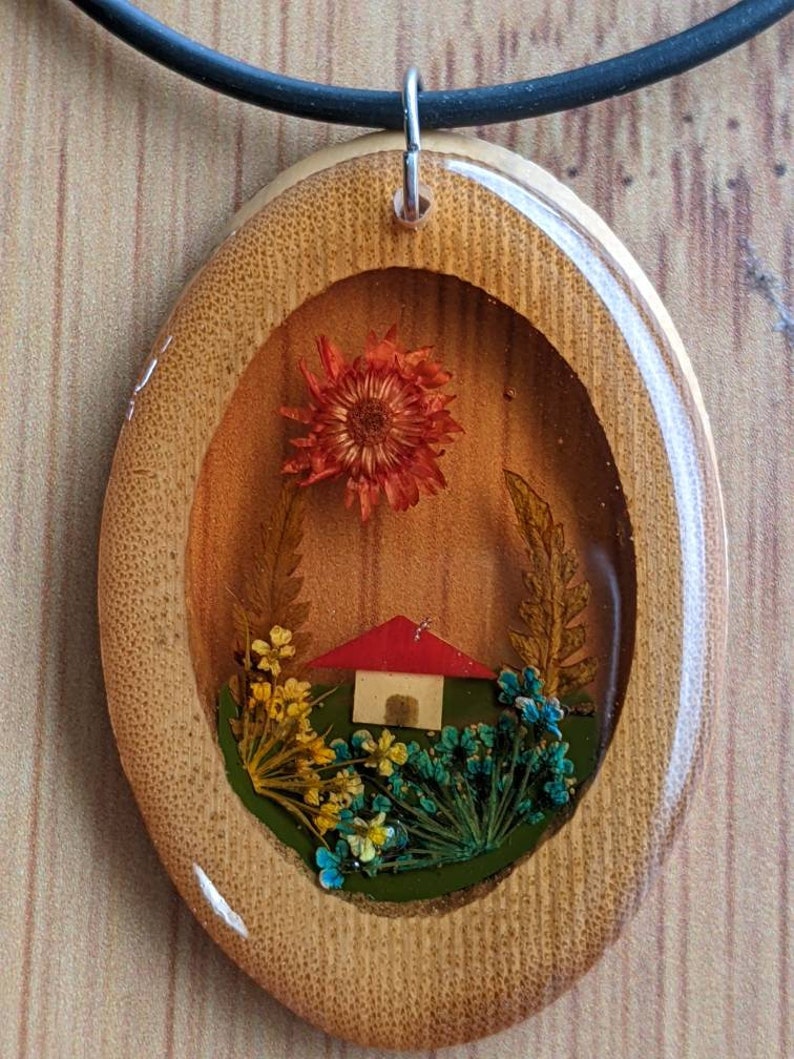 May include: A wooden oval pendant necklace with a clear resin insert featuring a miniature house and pressed flowers. The house has a red roof and white walls. The flowers are in shades of orange, yellow, blue, and green.