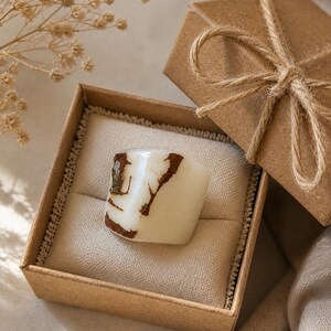 May include: A white and brown ring is displayed in an open brown gift box with a beige interior. The ring has a unique, organic shape with brown veining. The box is tied with twine, and a sprig of dried flowers is in the background.