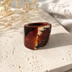 May include: A small, handcrafted ceramic ring holder. The holder is a rich brown with white and dark brown marbling. The ring holder is on a textured, off-white surface, with dried flowers in the background.