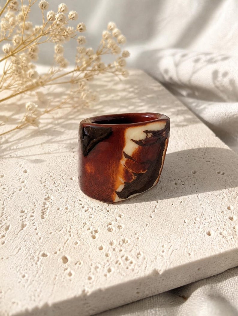 May include: A small, handcrafted ceramic ring holder. The holder is a rich brown with white and dark brown marbling. The ring holder is on a textured, off-white surface, with dried flowers in the background.