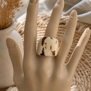 May include: A chunky, square ring with a cream-colored base and irregular brown marbling. The ring is displayed on a mannequin hand, showcasing its bold design. The ring's unique pattern and shape make it a statement piece.