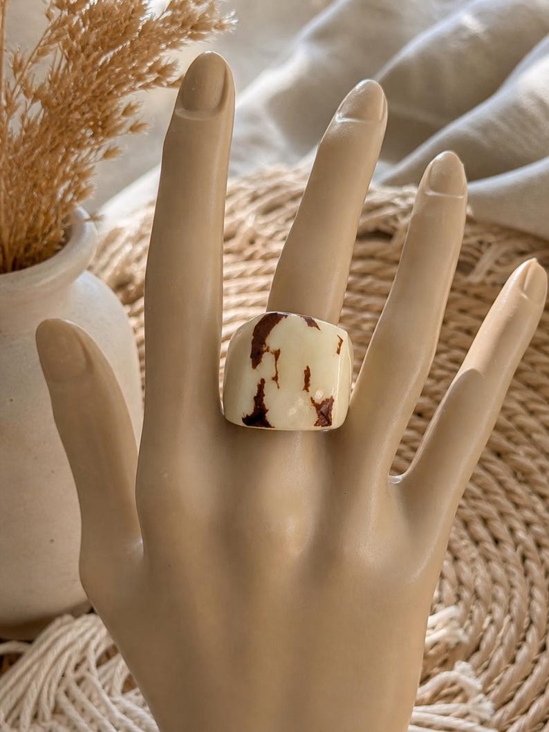 May include: A chunky, square ring with a cream-colored base and irregular brown marbling. The ring is displayed on a mannequin hand, showcasing its bold design. The ring's unique pattern and shape make it a statement piece.
