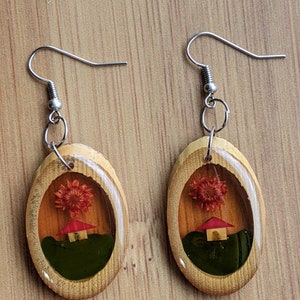 May include: A pair of oval-shaped wooden earrings with a clear resin insert. The resin contains a small red house and a dried red flower. The earrings have silver hooks.
