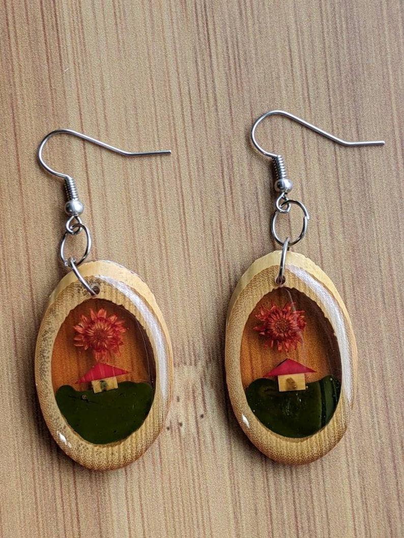 May include: A pair of oval-shaped wooden earrings with a clear resin insert. The resin contains a small red house and a dried red flower. The earrings have silver hooks.