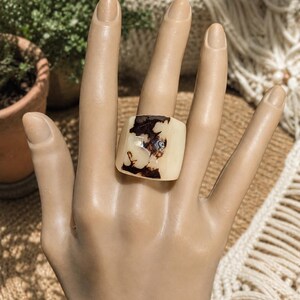 May include: A square, cream-colored ring with a dark brown, marbled design. The ring is worn on a mannequin hand, showcasing its unique pattern and shape. The ring's design is reminiscent of natural stone.