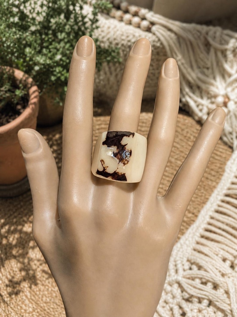 May include: A square, cream-colored ring with a dark brown, marbled design. The ring is worn on a mannequin hand, showcasing its unique pattern and shape. The ring's design is reminiscent of natural stone.