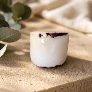 May include: A small, white ceramic cup with a rounded shape and a dark brown rim. The cup is set on a light-colored, textured surface, with soft greenery in the background. The cup's design is simple and minimalist.