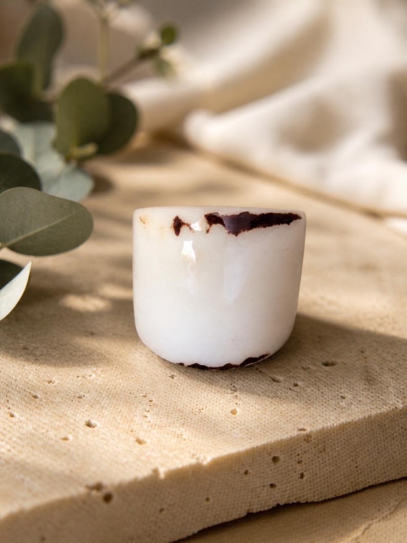 May include: A small, white ceramic cup with a rounded shape and a dark brown rim. The cup is set on a light-colored, textured surface, with soft greenery in the background. The cup's design is simple and minimalist.