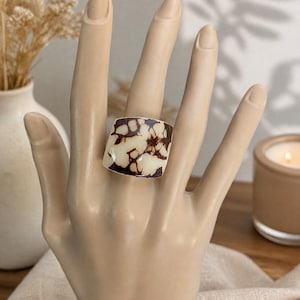 May include: A square ring with a cream-colored base and brown, organic-shaped patterns. The ring is displayed on a mannequin hand. The background includes a vase with dried flowers and a lit candle.
