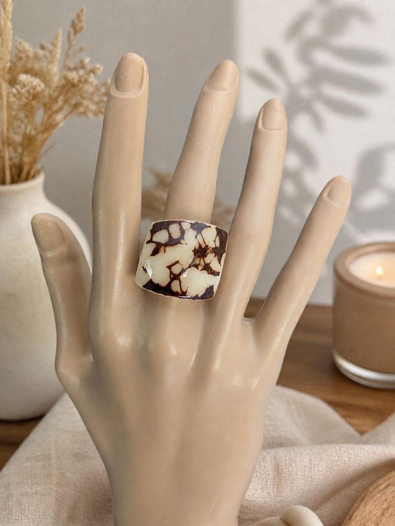 May include: A square ring with a cream-colored base and brown, organic-shaped patterns. The ring is displayed on a mannequin hand. The background includes a vase with dried flowers and a lit candle.
