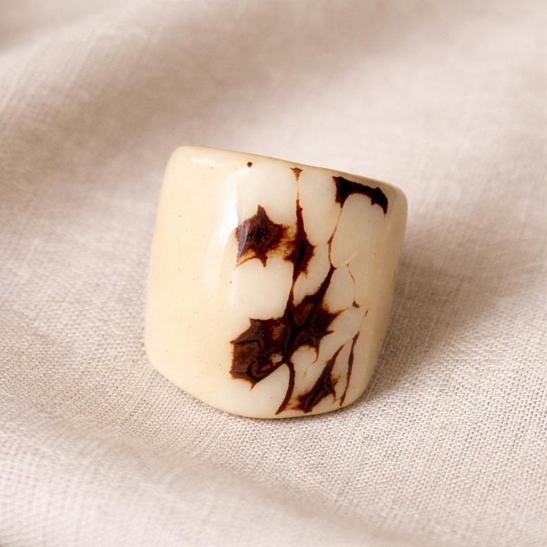 May include: A chunky, cream-colored ring with a glossy finish. The ring features a unique pattern of dark brown, organic shapes across its surface. The ring is a bold statement piece, perfect for adding a touch of artistic flair to any outfit.