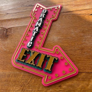 May include: A pink arrow-shaped sign with the word "EXIT" in gold lettering and the word "DRAMATIC" stacked vertically. The sign has a gold border and small gold circles around the edges. The sign is made of layered materials.