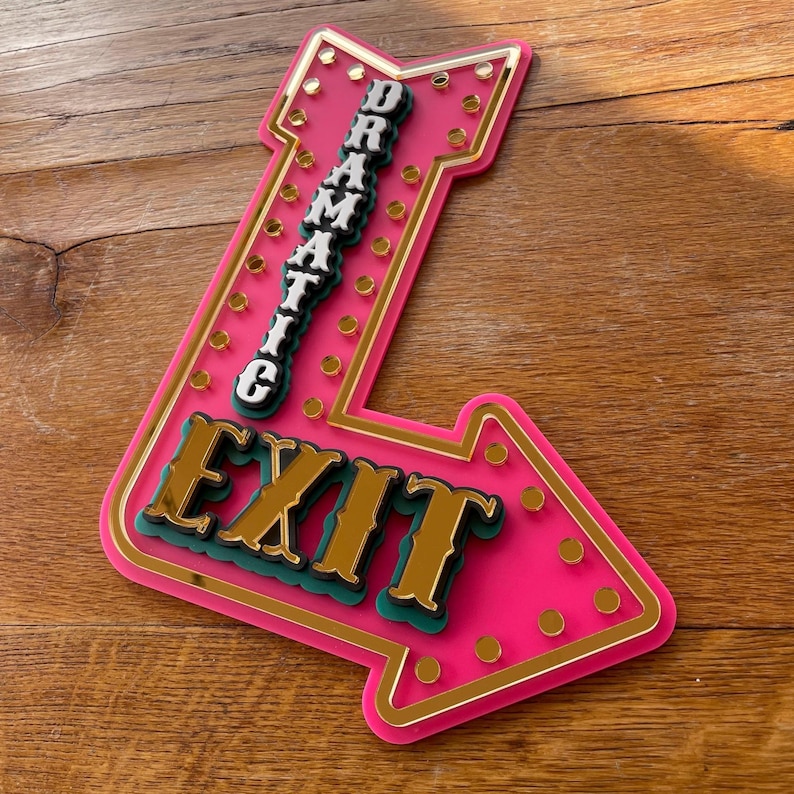 May include: A pink arrow-shaped sign with the word "EXIT" in gold lettering and the word "DRAMATIC" stacked vertically. The sign has a gold border and small gold circles around the edges. The sign is made of layered materials.