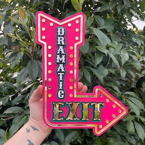 May include: A vibrant pink arrow-shaped sign with the words "DRAMATIC" and "EXIT" in gold lettering. The sign has a gold border and small white lights. The background is a lush green foliage.