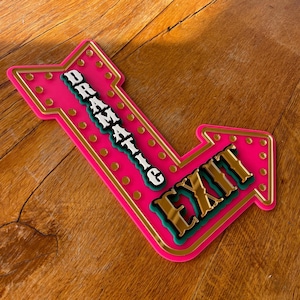 May include: A pink arrow-shaped sign with the words "DRAMATIC EXIT" in gold, white, and teal lettering. The sign has a gold border and small gold circles. The sign is on a wooden surface.