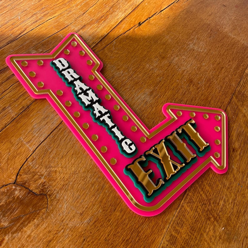May include: A pink arrow-shaped sign with the words "DRAMATIC EXIT" in gold, white, and teal lettering. The sign has a gold border and small gold circles. The sign is on a wooden surface.