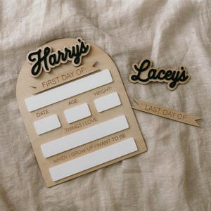 Op de afbeelding: Een houten bord met de naam "Harry's" en de tekst "Eerste dag van:" met ruimte om de datum, leeftijd, lengte, dingen die ze leuk vinden en wat ze willen worden als ze groot zijn te schrijven. Een apart houten bord met de naam "Lacey's" en de tekst "Laatste dag van:" is ook inbegrepen.