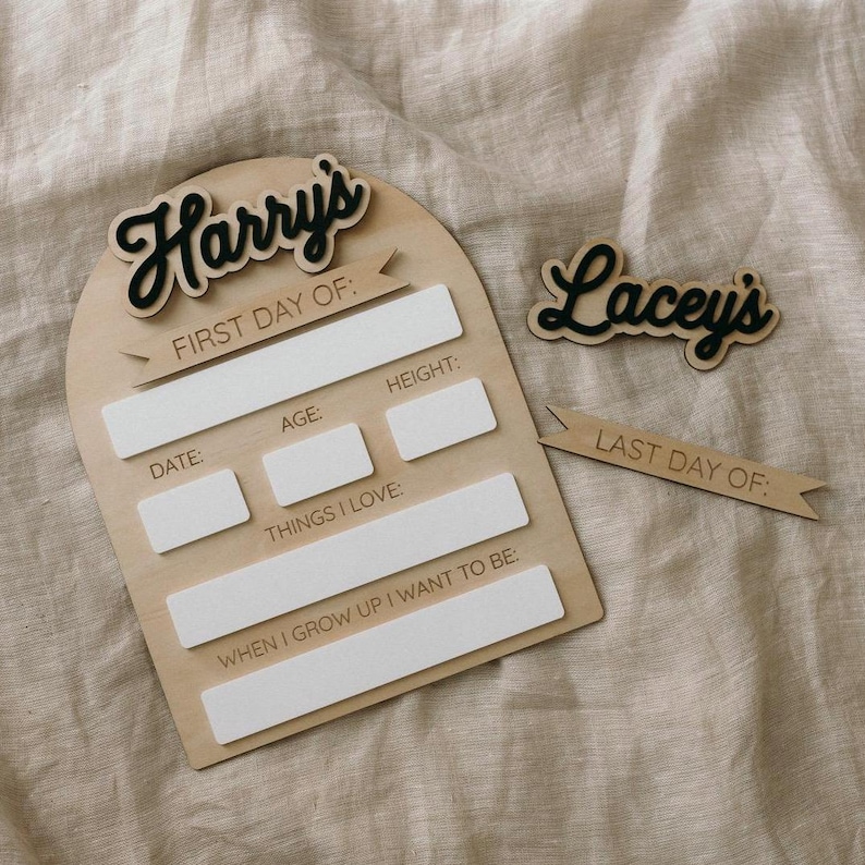 Op de afbeelding: Een houten bord met de naam "Harry's" en de tekst "Eerste dag van:" met ruimte om de datum, leeftijd, lengte, dingen die ze leuk vinden en wat ze willen worden als ze groot zijn te schrijven. Een apart houten bord met de naam "Lacey's" en de tekst "Laatste dag van:" is ook inbegrepen.