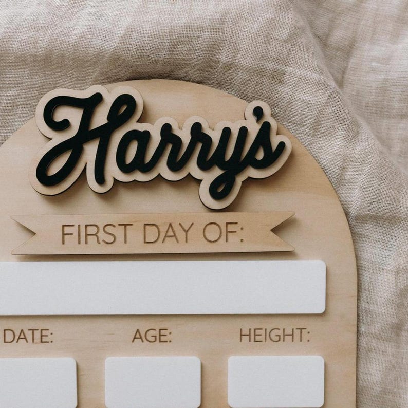 Op de afbeelding: Een houten bord met de naam "Harry's" in zwarte cursieve letters. Het bord heeft een banner met de tekst "Eerste dag van:" en ruimte om data, leeftijd en lengte te noteren.