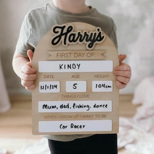 Op de afbeelding: Een houten bord met een afgeronde bovenkant en een zwarte inscriptie van "Harry's" in een cursief schrift. Het bord is versierd met een wit banner met de tekst "First Day Of:". Onder het banner bevinden zich witte labels met zwarte tekst, waaronder "&Kappa;&Iota;&Nu;&Omicron;&Upsilon;", "Datum: 31/1/24", "Leeftijd: 5", "Lengte: 104 cm", "Dingen die ik leuk vind: Mama, papa, vissen, dansen" en "Wanneer ik groot ben, wil ik zijn: Autocoureur".