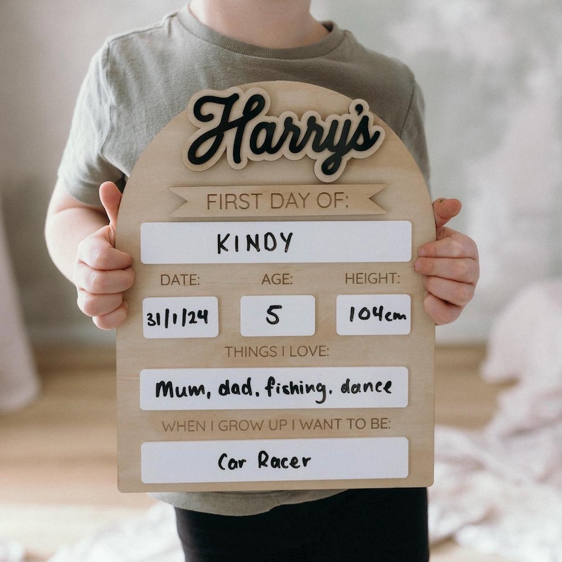 Op de afbeelding: Een houten bord met een afgeronde bovenkant en een zwarte inscriptie van "Harry's" in een cursief schrift. Het bord is versierd met een wit banner met de tekst "First Day Of:". Onder het banner bevinden zich witte labels met zwarte tekst, waaronder "&Kappa;&Iota;&Nu;&Omicron;&Upsilon;", "Datum: 31/1/24", "Leeftijd: 5", "Lengte: 104 cm", "Dingen die ik leuk vind: Mama, papa, vissen, dansen" en "Wanneer ik groot ben, wil ik zijn: Autocoureur".