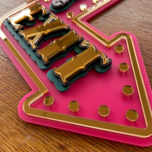 May include: A pink arrow-shaped sign with gold mirrored accents and lettering. The sign has a gold border and gold circular embellishments. The letters are gold and sit on green and black bases.