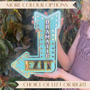 May include: A light blue arrow-shaped sign with a metallic gold border and gold accents. The sign features the vertical text "DRAMATIC" and the horizontal text "EXIT" in a bold, dark font. The design includes circular cutouts along the edges.