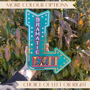 May include: A teal and gold-coloured arrow-shaped sign with the word "DRAMATIC" vertically and "EXIT" horizontally. The sign has a gold border and is decorated with small, round, white dots. The text "MORE COLOUR OPTIONS" and "CHOICE OF LEFT OR RIGHT" are also visible.