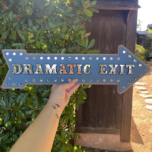 May include: A blue arrow-shaped sign with a metallic border and silver circular accents along the edges. The sign features the text "DRAMATIC EXIT" in large, reflective, silver-toned capital letters centered across the middle of the arrow.