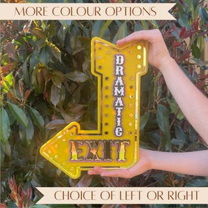 May include: A yellow arrow-shaped sign with the word "DRAMATIC" vertically and "EXIT" horizontally. The sign has a gold border and is held up against a background of green leaves. The text "MORE COLOUR OPTIONS" and "CHOICE OF LEFT OR RIGHT" are also visible.